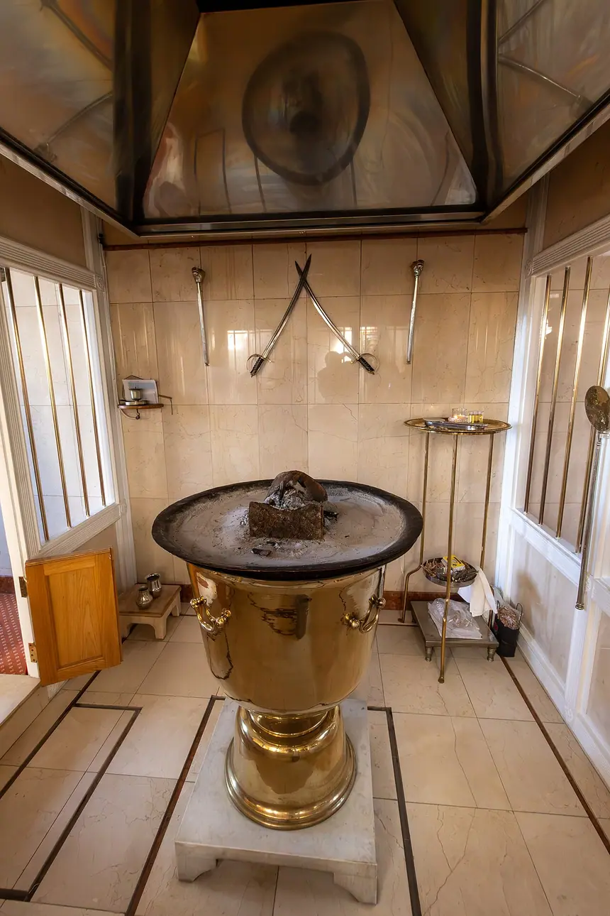 Phòng lửa trong Trung tâm Zoroastrian ở Rayners Lane, London: bình lửa kim loại (afarganyu) đặt trên bệ đá giữa phòng lát gạch trắng. Trung tâm nằm trong một rạp chiếu phim cũ, phòng lửa ở chỗ buồng máy chiếu ngày trước. Ảnh chụp tháng 9 năm 2023.