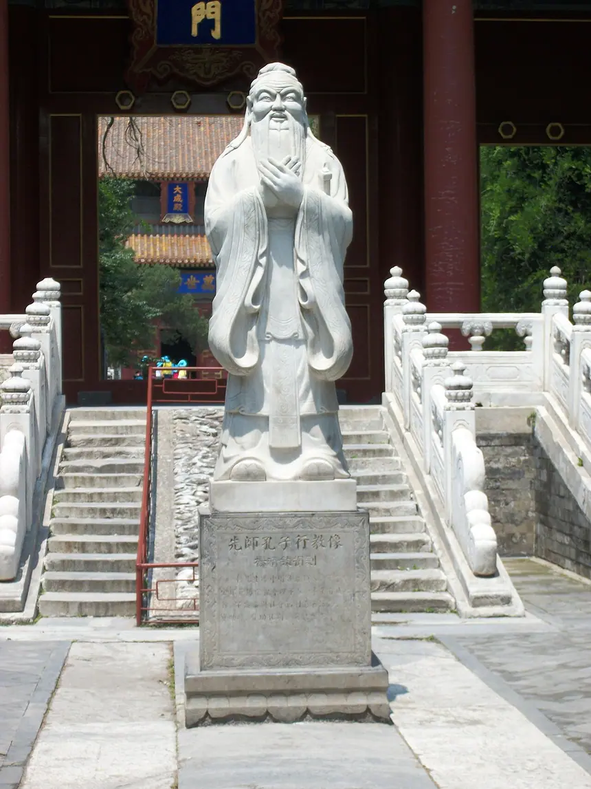 Tượng đá Khổng Tử đứng chắp tay ngoài sân Khổng miếu Bắc Kinh, làm theo dáng bức "hành giáo". Tượng mới. Bệ khắc chữ "Tiên sư Khổng Tử hành giáo tượng".