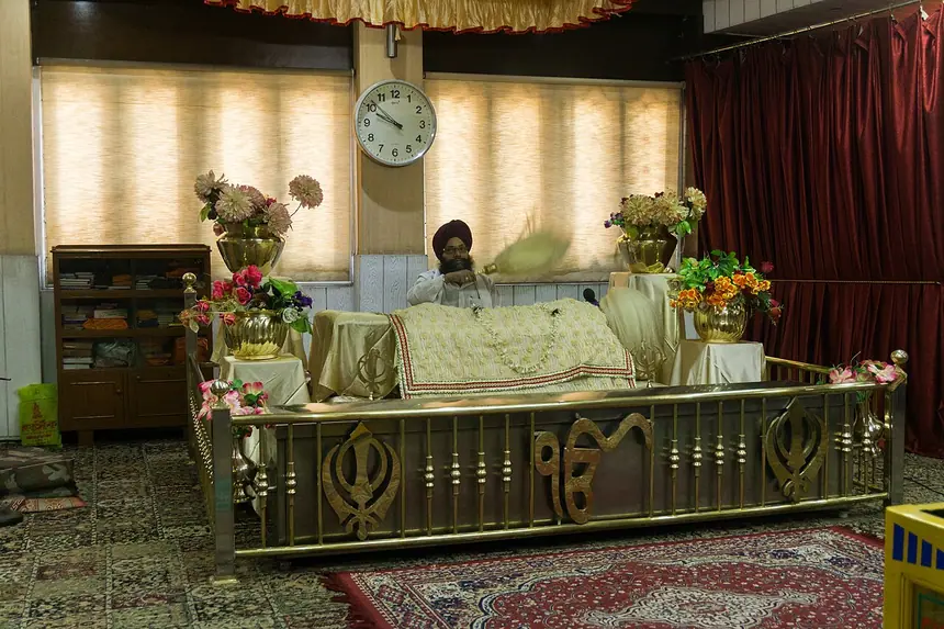 Guru Granth Sahib an vị trên bục trong Gurdwara Bara Sikh Sangat: Kinh phủ khăn rumala, vị granthi ngồi phía sau, lan can mang hình Khanda và chữ Ik Onkar. Ảnh chụp năm 2017.