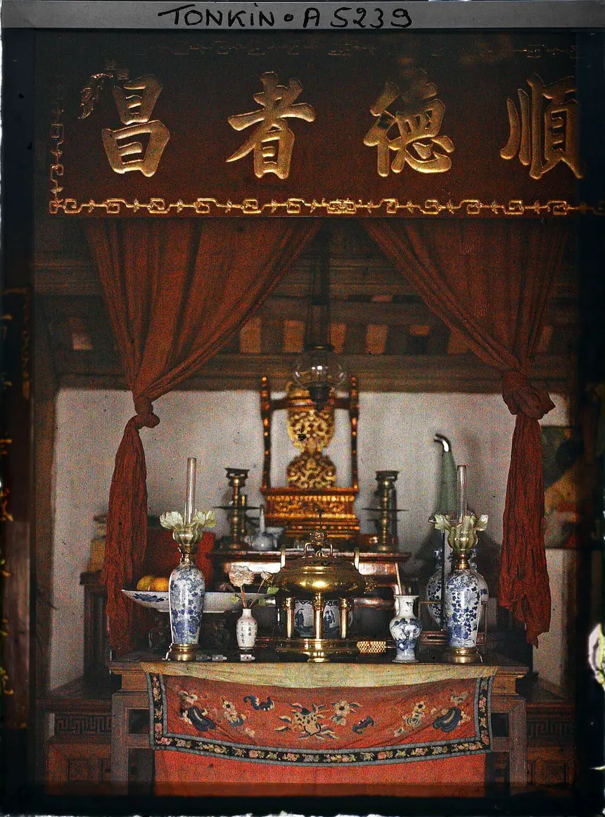 Bàn thờ tổ tiên trong một nhà khá giả ở Hà Nội, ảnh màu autochrome của Léon Busy, năm 1914 tới 1915. Trên cao là bức hoành phi bốn chữ Hán, dưới là màn đỏ vén hai bên, trong cùng có chiếc ngai thếp vàng, phía trước là đỉnh đồng, chân nến và lọ hoa. Lưu trữ Albert Kahn.