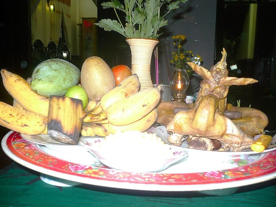 Midnight outdoor Lunar New Year’s Eve offering table. Open-air courtyard table prepared at midnight for the incoming Year Commissioner, featuring a boiled rooster, five-fruit tray, yellow chrysanthemums, sticky rice and a lit oil lamp.
