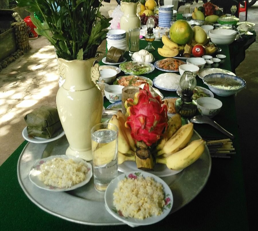 Pure vegetarian offering tray for Full Moon and New Moon rites. Modest household vegetarian offering featuring mung-bean sticky rice, leaf-wrapped cakes, plant-based savory dishes, dragon fruit and ripe bananas, a lit oil lamp and fresh flowers.